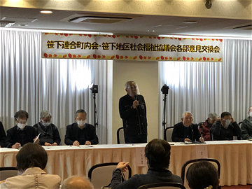 笹下連合町内会・笹下地区社会福祉協議会各部意見交換会の横断幕の前に立って挨拶する会長の写真